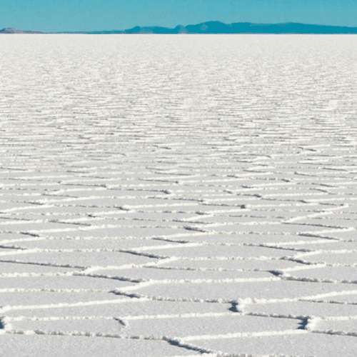 A panoramic view of a vast salt flat, characterized by a bright white surface with intricate, geometric patterns formed by salt crystals. The horizon features distant mountains and a clear blue sky.